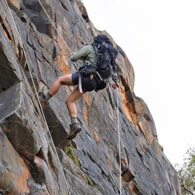 Evan abseiling down a dark rock face with a big pack on his back.