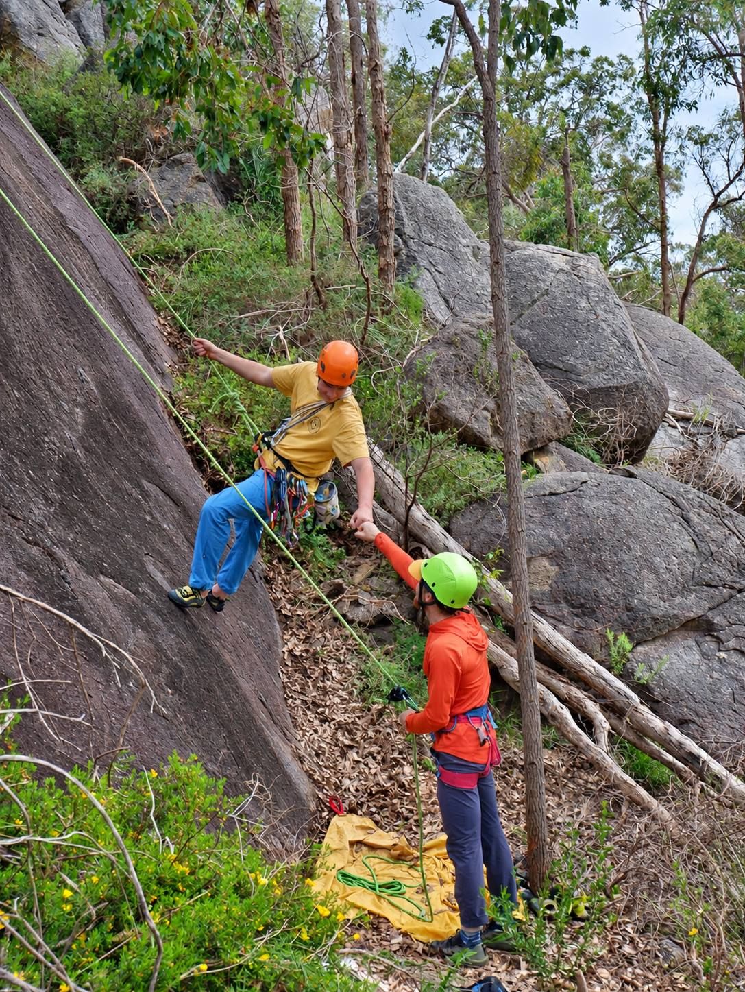 A climber in an orange helmet starting up a granite slab on a top rope, while a belayer in a green helmet holds the rope below.